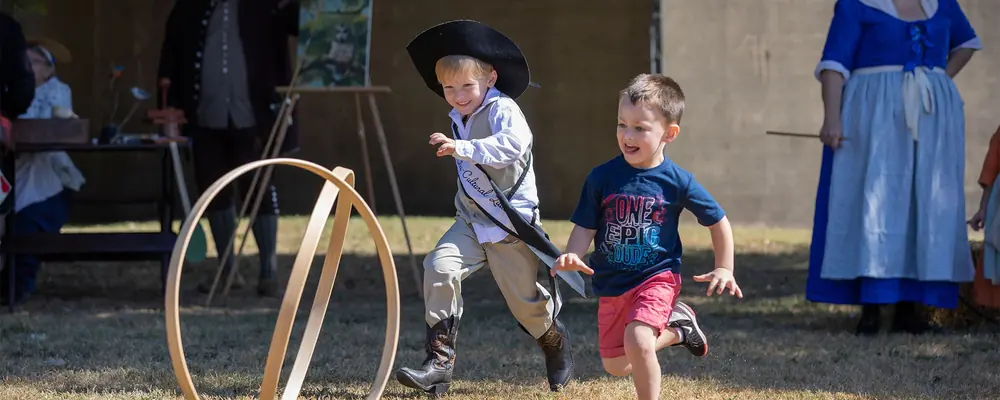 Two children racing a wooden hoop at a living-history event, with costumed interpreters behind them